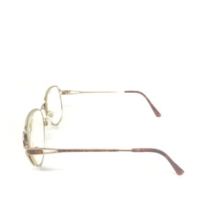 K&ouml;nnte beinhalten: Eine Brille mit einem goldfarbenen Metallrahmen und ovalen Gl&auml;sern. Die B&uuml;gel sind dunkelbraun. Die Brille ist vor einem wei&szlig;en Hintergrund platziert.