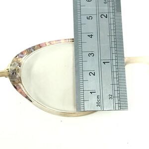 May include: A silver-toned, oval-shaped object with a white interior is measured with a ruler. The object is approximately 3 inches long. The rim of the object has a decorative, iridescent pattern.