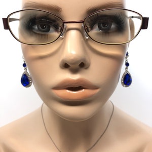 May include: A mannequin head wearing eyeglasses with a burgundy frame and teardrop earrings with large blue gems and silver accents. A silver necklace is also visible.