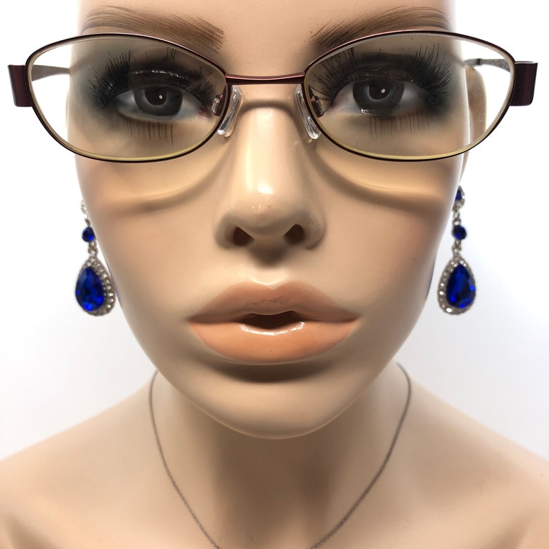 May include: A mannequin head wearing eyeglasses with a burgundy frame and teardrop earrings with large blue gems and silver accents. A silver necklace is also visible.