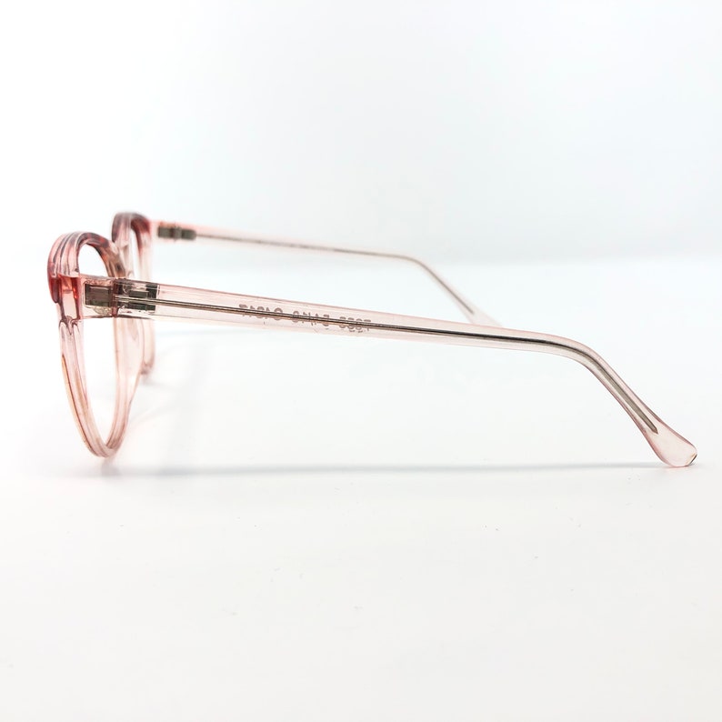 May include: Pale pink eyeglasses with a rounded frame and thin temples. The glasses have a transparent frame and arms, with the text "FABIO CONTI 3227" printed on the temple. The glasses are set against a white background.