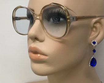 May include: A mannequin wearing large, round, clear, plastic framed glasses with blue lenses. The mannequin is wearing blue teardrop-shaped earrings with rhinestones.