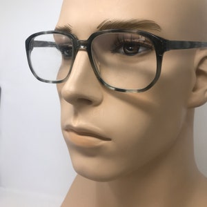 Puede incluir: Gafas de montura cuadrada con un patr&oacute;n moteado gris y blanco. Las gafas tienen lentes transparentes y se exhiben en un maniqu&iacute;. Las monturas son gruesas y el estilo general es vintage.