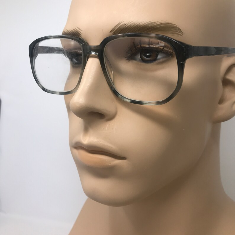 Puede incluir: Gafas de montura cuadrada con un patr&oacute;n moteado gris y blanco. Las gafas tienen lentes transparentes y se exhiben en un maniqu&iacute;. Las monturas son gruesas y el estilo general es vintage.