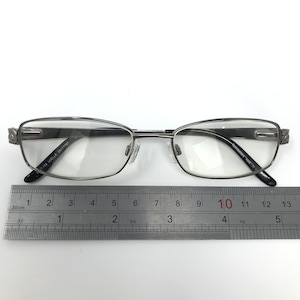May include: Silver-framed eyeglasses with clear lenses. The glasses have a rectangular shape with rounded edges and black temple tips. A ruler is placed below the glasses for measurement.