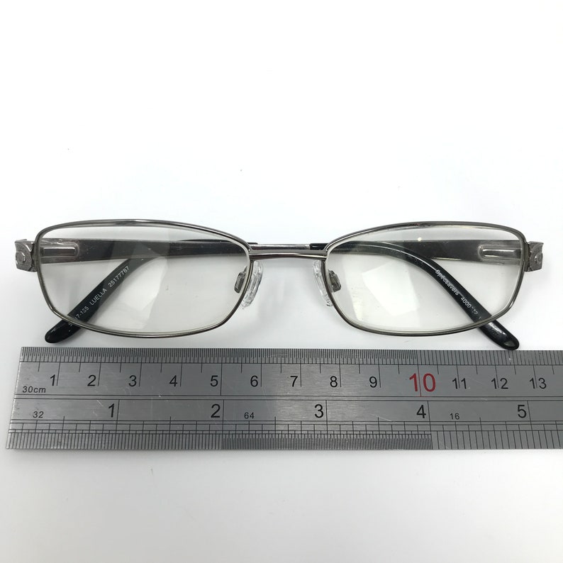 May include: Silver-framed eyeglasses with clear lenses. The glasses have a rectangular shape with rounded edges and black temple tips. A ruler is placed below the glasses for measurement.