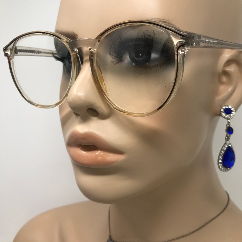Puede incluir: Gafas transparentes de montura redonda con un tinte claro. Las gafas est&aacute;n colocadas en una cabeza de maniqu&iacute;, con una vista de cerca de la cara. El maniqu&iacute; tambi&eacute;n lleva pendientes colgantes azules y plateados.