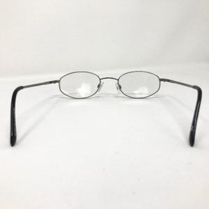 Op de afbeelding: Ovale bril met een donkergrijs metalen montuur en heldere glazen. De pootjes zijn zwart en steken naar buiten. De bril staat tegen een witte achtergrond.