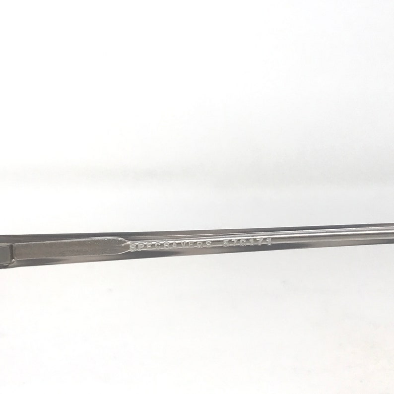 May include: Close-up of a gray eyeglass frame arm. The text "SPECSAVERS 570474" is printed on the arm. The frame is a neutral color, suitable for various styles.