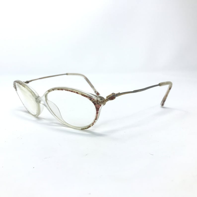 May include: Clear plastic oval-shaped eyeglasses with a gold metal frame and decorative accents. The frame has a subtle floral pattern.