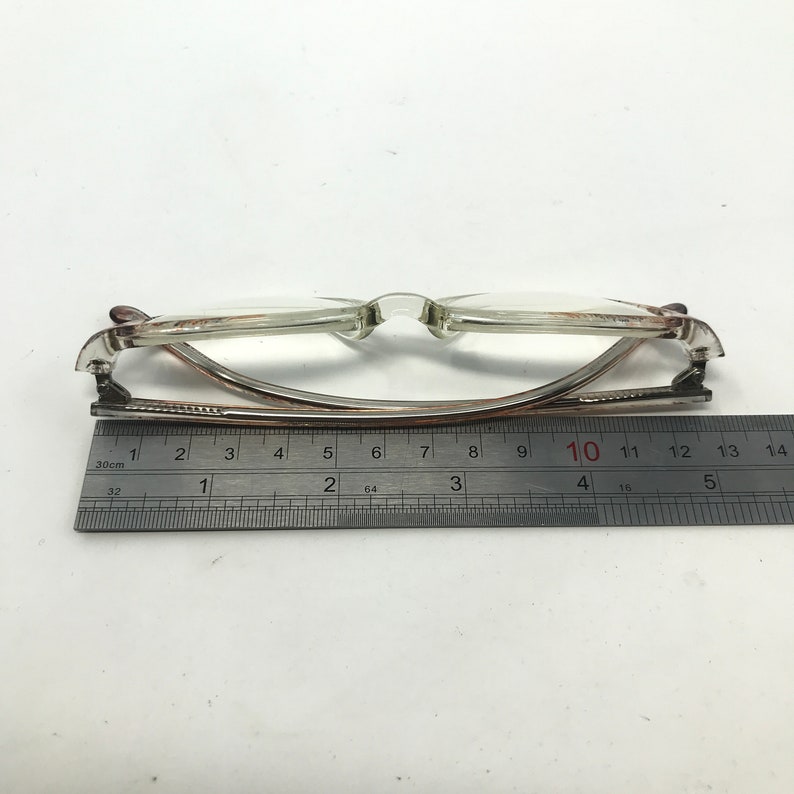 May include: Transparent-framed spectacles with a metal and plastic construction. The glasses are measured with a ruler, showing a length of approximately 13 cm. The lenses are clear, and the frame has a subtle brown tint.