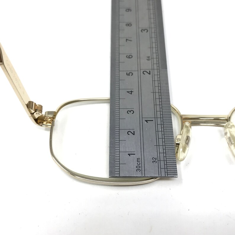 May include: Gold-tone metal eyeglasses with a hinge detail. The glasses are measured against a ruler, showing the frame width is approximately 1 1/2 inches.