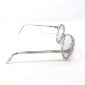 May include: Clear, round-framed eyeglasses with a light tint. The frames and arms are a translucent, pale pink color. The glasses are set against a white background.