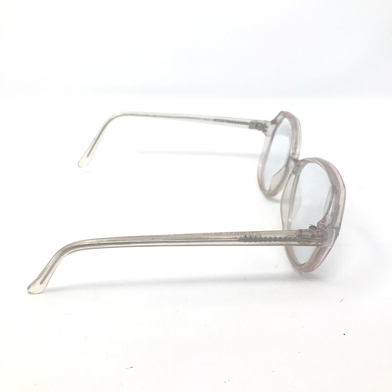 May include: Clear, round-framed eyeglasses with a light tint. The frames and arms are a translucent, pale pink color. The glasses are set against a white background.