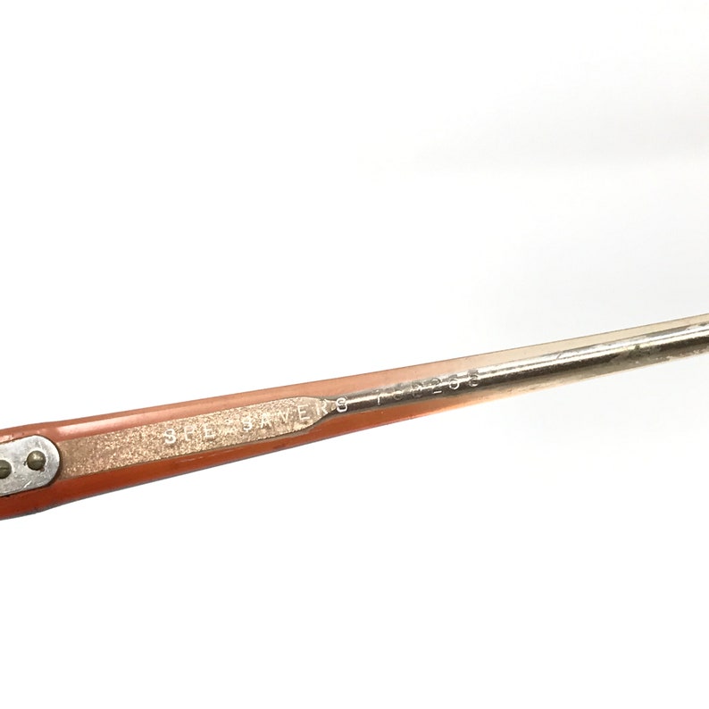 May include: Close-up of a vintage eyeglasses arm. The arm is a light brown color with the text "SHE SAVERS" and "T 50203" printed on it. The arm has a silver metal hinge.
