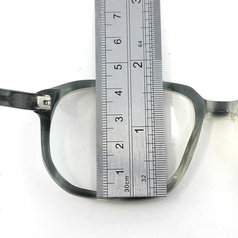 May include: Close-up of a pair of gray eyeglasses with a ruler placed over the lens. The ruler shows a measurement of approximately 1 inch. The glasses have a rectangular frame and clear lenses.