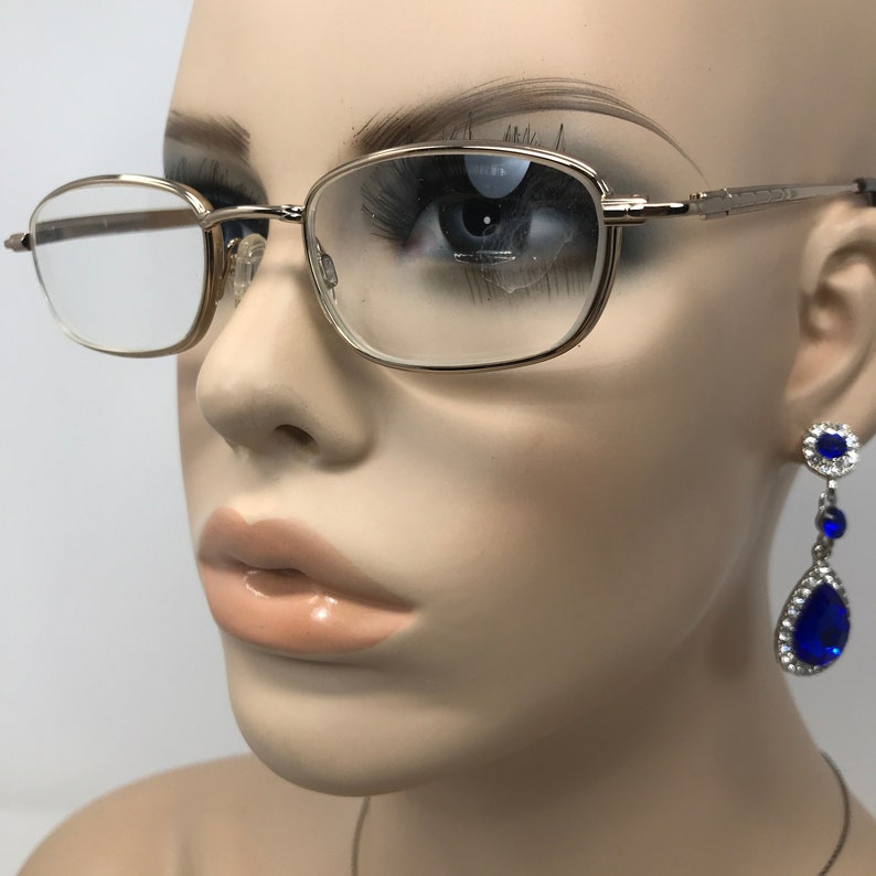 May include: Gold-tone rectangular eyeglasses with clear lenses. The glasses are on a mannequin. The mannequin is also wearing a pair of blue and silver drop earrings. The glasses have a thin frame and a classic design.