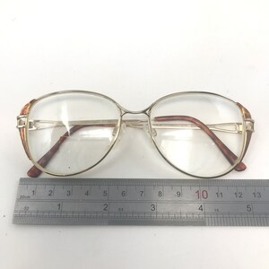 K&ouml;nnte beinhalten: Goldgerahmte Brille mit klaren Gl&auml;sern. Die Fassungen haben eine rechteckige Form mit braunen Akzenten an den B&uuml;geln. Ein Lineal mit Zentimetermessung liegt unter der Brille.