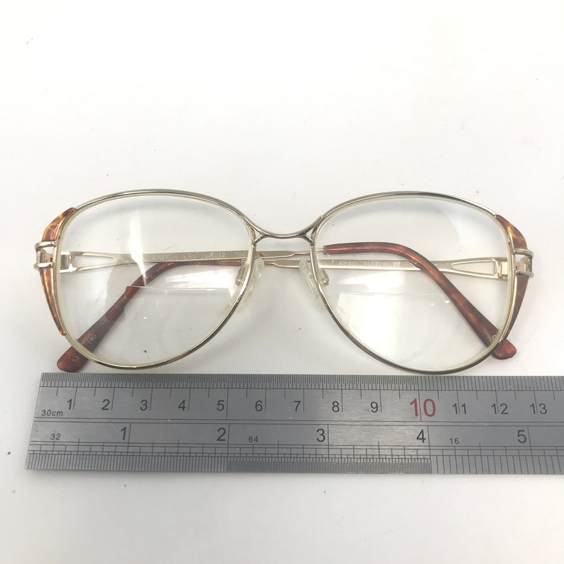 K&ouml;nnte beinhalten: Goldgerahmte Brille mit klaren Gl&auml;sern. Die Fassungen haben eine rechteckige Form mit braunen Akzenten an den B&uuml;geln. Ein Lineal mit Zentimetermessung liegt unter der Brille.