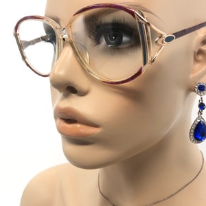 K&ouml;nnte beinhalten: Brille mit transparentem Rahmen, goldfarbenem Akzent und lila Zierleiste. Die Brille wird mit einem h&auml;ngenden Ohrring kombiniert, der einen gro&szlig;en, tropfenf&ouml;rmigen blauen Edelstein umgeben von kleineren, klaren Steinen aufweist. Der Ohrring ist silberfarben.
