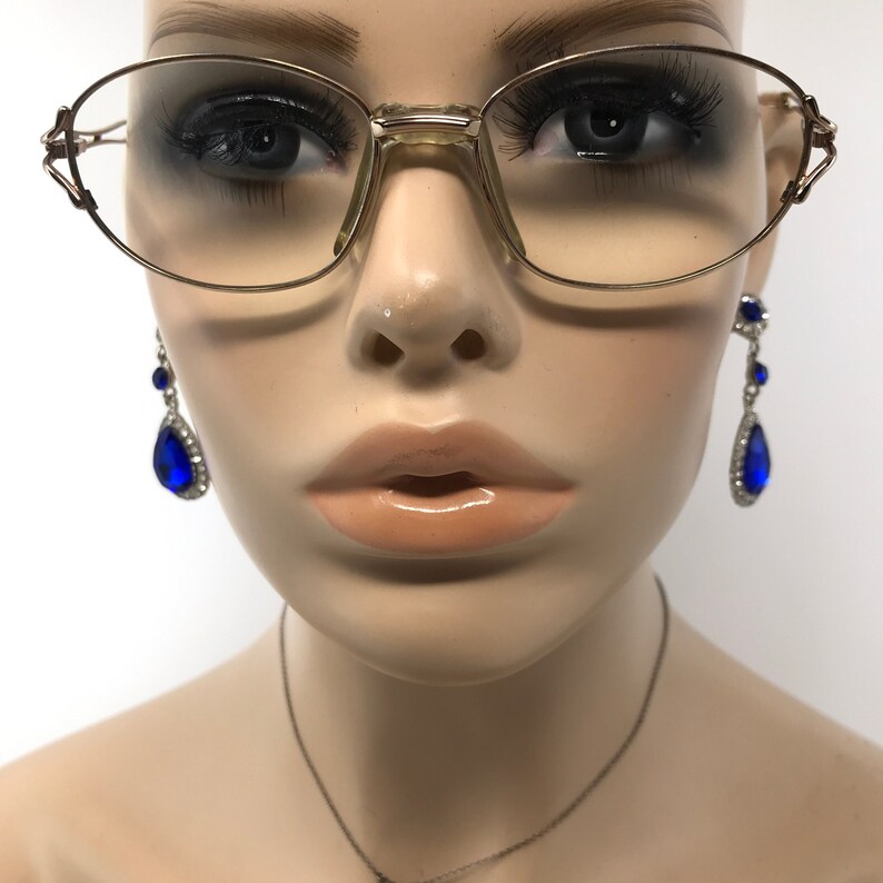Peut inclure: Gros plan d'un mannequin portant des lunettes ovales &agrave; monture dor&eacute;e et des boucles d'oreilles en forme de larme bleues avec des strass.