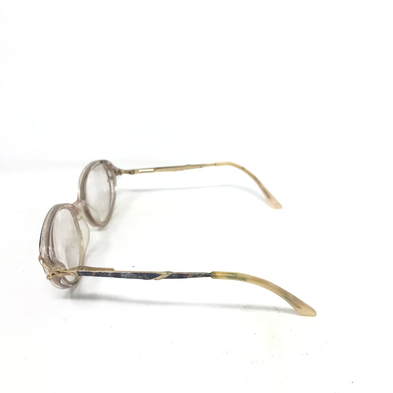 May include: A pair of round-framed eyeglasses with clear lenses. The frames are a light, translucent color with gold-toned metal arms and temples. The glasses are set against a white background.