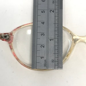 K&ouml;nnte beinhalten: Nahaufnahme einer Brille mit einem klaren Gestell und rosa Akzenten. Die Brille wird mit einem Lineal gemessen, wobei die Breite des Gestells etwa 3,8 cm betr&auml;gt.
