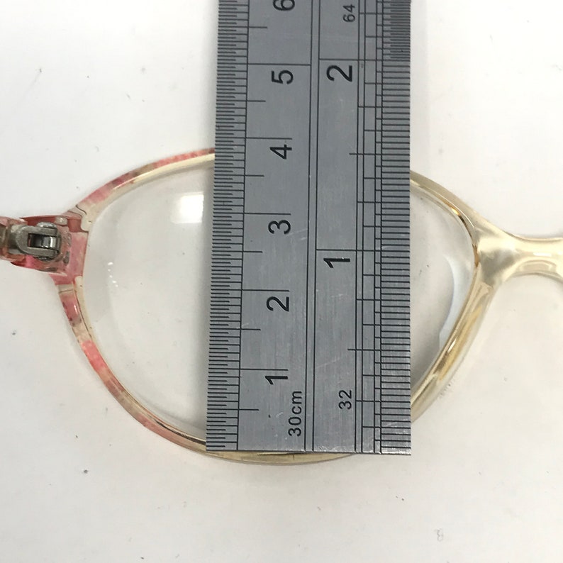 K&ouml;nnte beinhalten: Nahaufnahme einer Brille mit einem klaren Gestell und rosa Akzenten. Die Brille wird mit einem Lineal gemessen, wobei die Breite des Gestells etwa 3,8 cm betr&auml;gt.