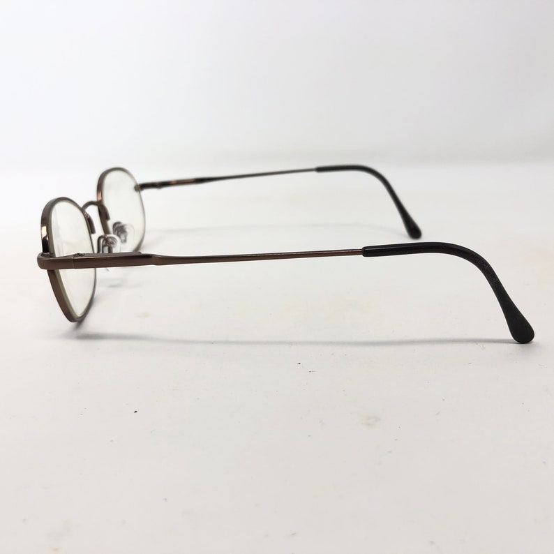 Puede incluir: Un par de gafas con una montura fina de metal color bronce y lentes redondas. Las patillas son de color marr&oacute;n oscuro y se curvan hacia la oreja. Las gafas est&aacute;n sobre un fondo blanco.