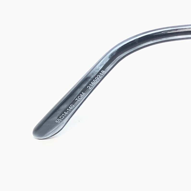 May include: Close-up of the temple of a pair of eyeglasses. The dark grey arm features the text "55▢14-140 TOM 21659218". The glasses have a sleek, modern design.