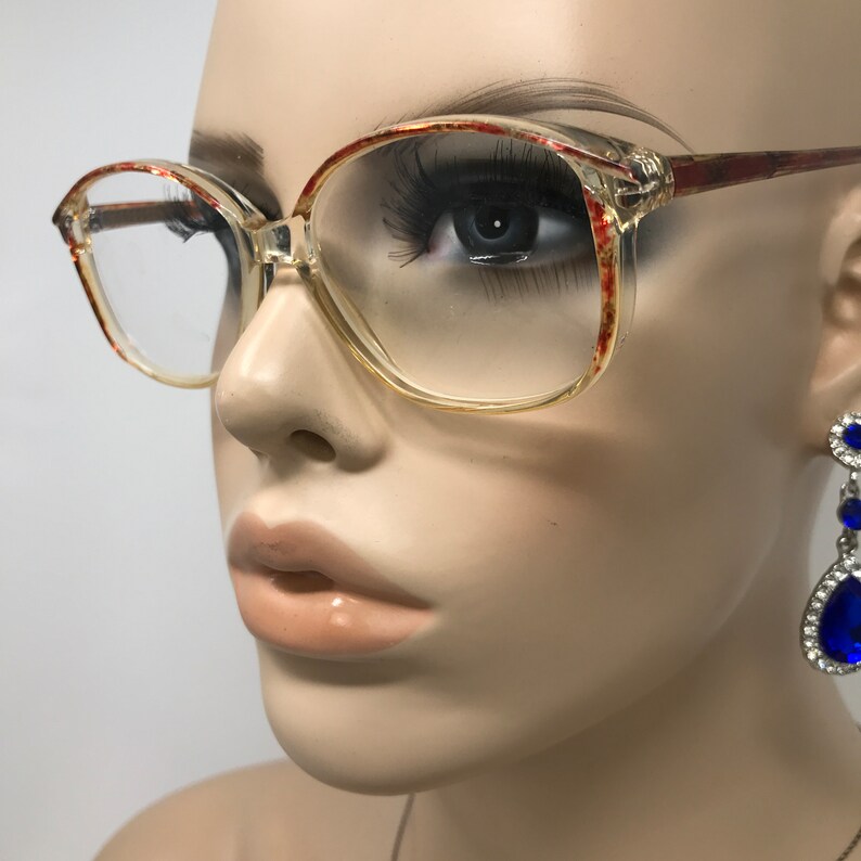 May include: Clear-framed eyeglasses with a square shape and red and gold accents on the upper rims and temples. The glasses are shown on a mannequin. A blue and silver earring is visible on the right side.