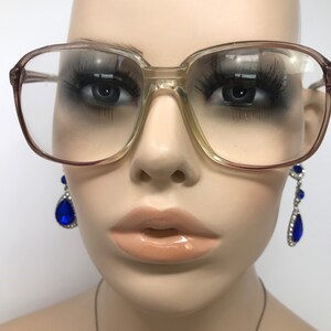 May include: Clear-lensed eyeglasses with a light brown frame. The glasses have a square shape and are set on a mannequin. The mannequin is wearing long, dark eyelashes and blue teardrop earrings.