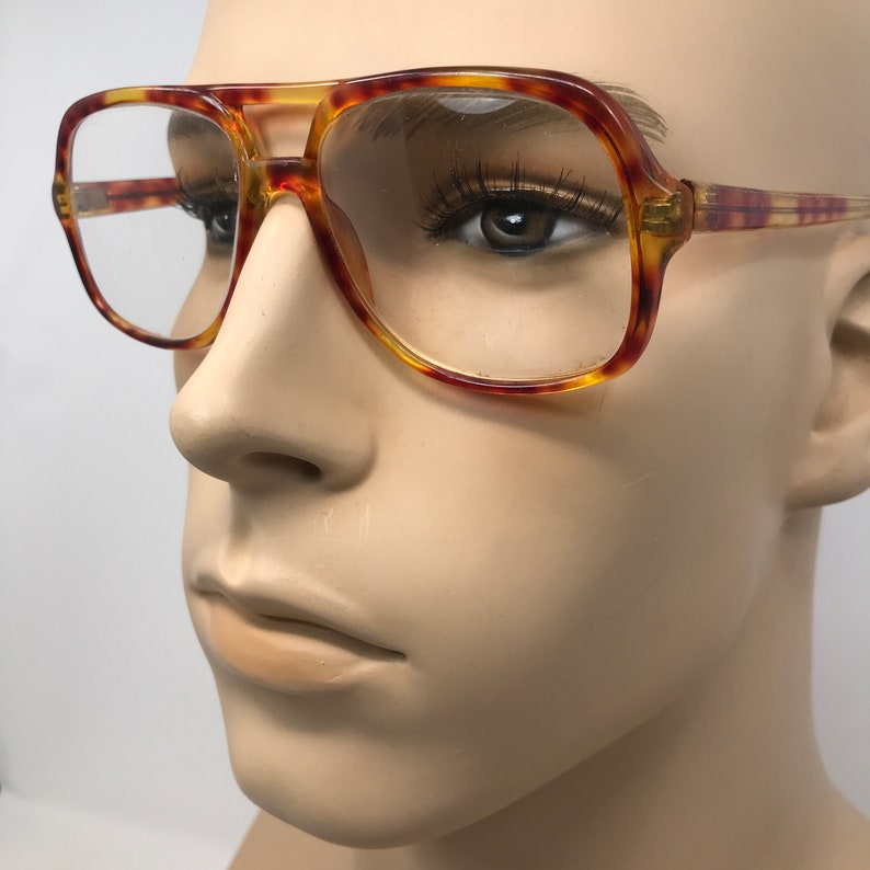 May include: Tortoiseshell-patterned eyeglasses with a square frame and a double bridge. The glasses have clear lenses and a classic, vintage design. The frames are a mix of amber and brown hues.