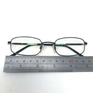 Puede incluir: Gafas rectangulares negras con lentes transparentes. Las gafas tienen una montura fina y oscura y est&aacute;n colocadas junto a una regla. La regla muestra medidas en cent&iacute;metros.