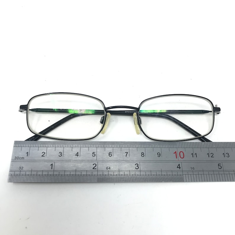 Puede incluir: Gafas rectangulares negras con lentes transparentes. Las gafas tienen una montura fina y oscura y est&aacute;n colocadas junto a una regla. La regla muestra medidas en cent&iacute;metros.
