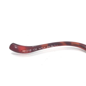 May include: Close-up of the brown plastic arm of a pair of glasses with the text "50□18-135 NOLA 24LBE-7-1" printed on it.