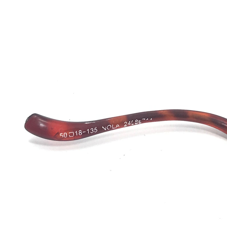 May include: Close-up of the brown plastic arm of a pair of glasses with the text "50□18-135 NOLA 24LBE-7-1" printed on it.