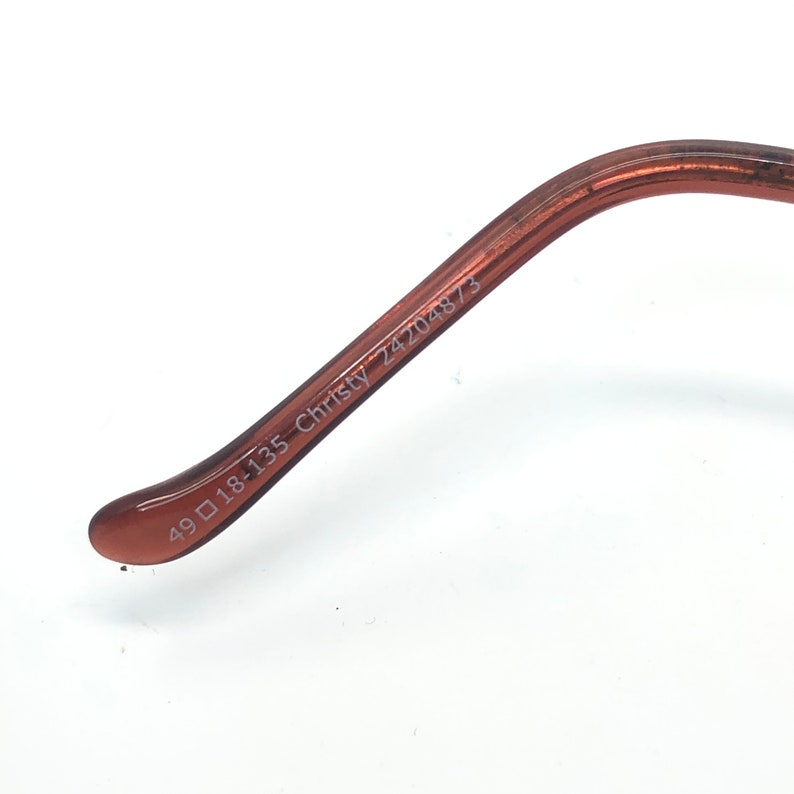 K&ouml;nnte beinhalten: Nahaufnahme des B&uuml;gels einer Brille. Der B&uuml;gel hat eine durchscheinende braune Farbe. Der Text "49 18-135 Christy 24204873" ist in Wei&szlig; auf dem B&uuml;gel aufgedruckt.