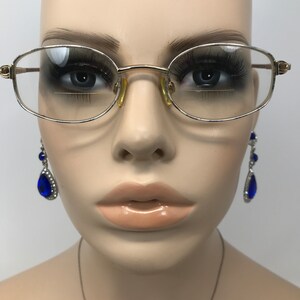 May include: Gold-rimmed spectacles with clear lenses. The glasses have a classic oval shape. The mannequin is also wearing blue teardrop earrings. The image is set against a neutral background.