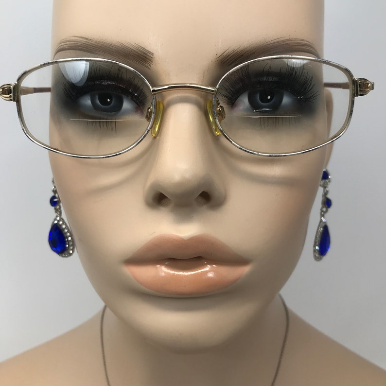 May include: Gold-rimmed spectacles with clear lenses. The glasses have a classic oval shape. The mannequin is also wearing blue teardrop earrings. The image is set against a neutral background.