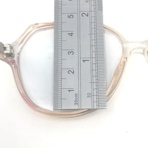May include: Clear-framed eyeglasses with a ruler placed over the lens. The ruler shows measurements in inches and centimeters. The glasses have a light pinkish hue and a unique hexagonal shape.