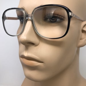 May include: Square-framed spectacles with a black and clear frame. The glasses have a thick, bold design with a clear lens. The temples are a light brown colour. The spectacles are on a mannequin.