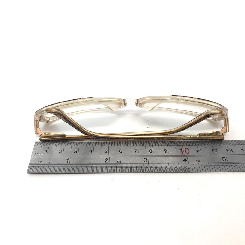 May include: A pair of gold-rimmed glasses with a brown tortoise shell pattern on the frame. The glasses are measured with a ruler, showing a length of 10 centimeters.