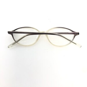 May include: Clear eyeglasses with a light gray frame and temples. The lenses are oval-shaped, and the temples have a slight curve. The glasses are set against a white background.