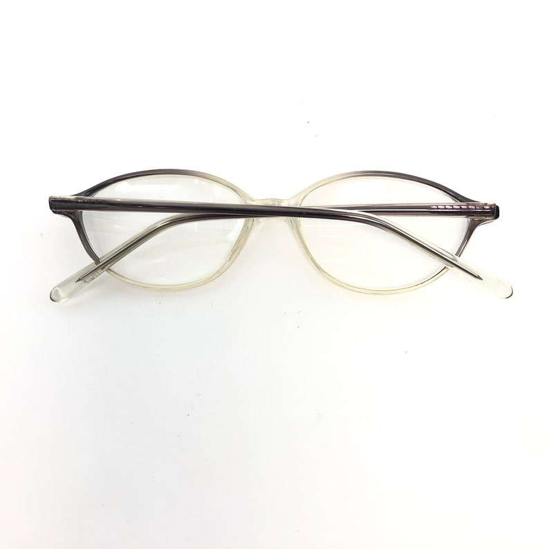 May include: Clear eyeglasses with a light gray frame and temples. The lenses are oval-shaped, and the temples have a slight curve. The glasses are set against a white background.