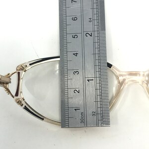 Puede incluir: Gafas con montura plateada y lentes transparentes. Se miden con una regla, mostrando un ancho de aproximadamente 3,2 cm. Las monturas tienen detalles en negro y burdeos. Fondo blanco.
