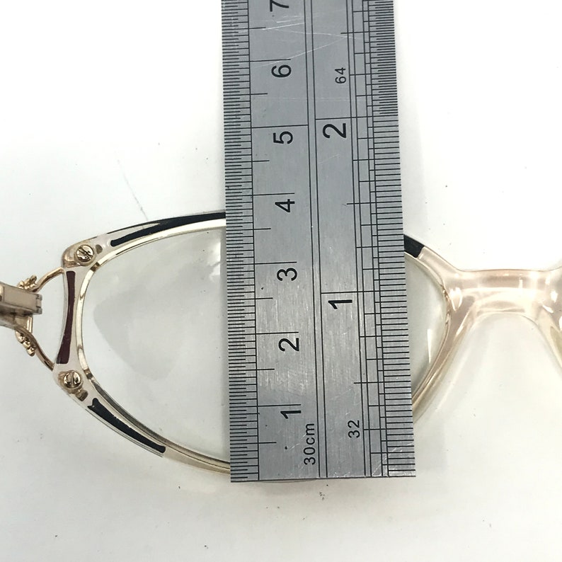 Puede incluir: Gafas con montura plateada y lentes transparentes. Se miden con una regla, mostrando un ancho de aproximadamente 3,2 cm. Las monturas tienen detalles en negro y burdeos. Fondo blanco.