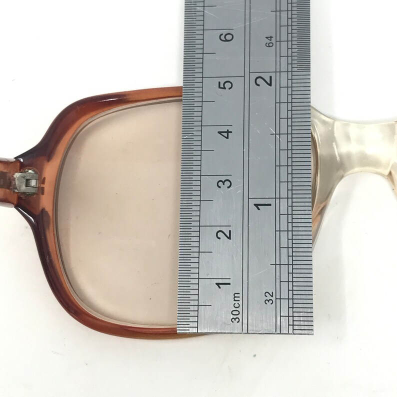 May include: Brown-framed eyeglasses with a rectangular lens shape. The frame is measured with a ruler, showing a lens width of approximately 2 inches. The glasses have a light tint and a classic design.