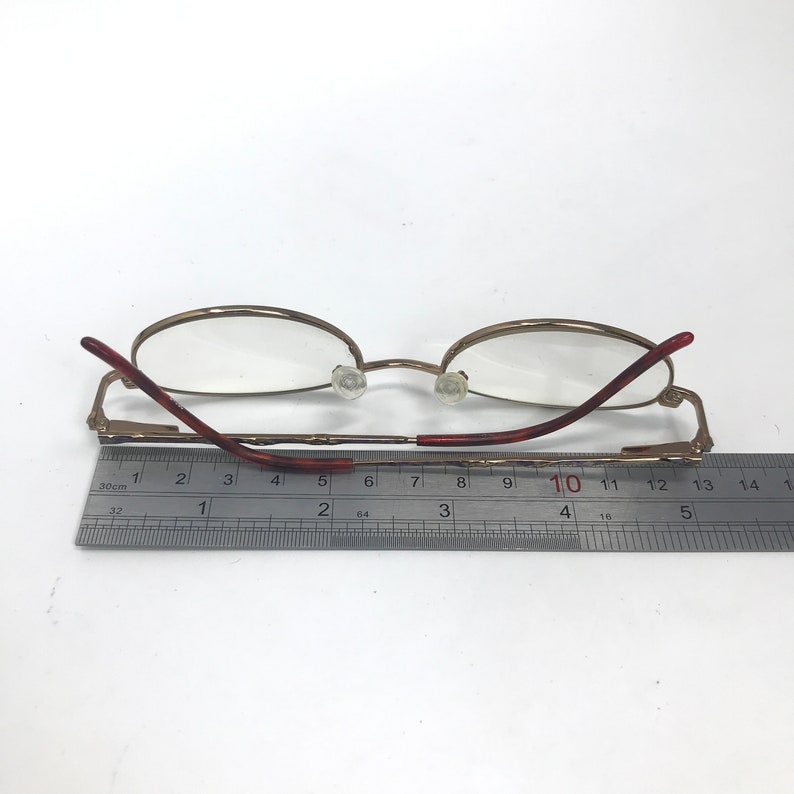 May include: Gold-tone metal eyeglasses with round lenses and brown plastic arms. The eyeglasses are measured with a ruler, showing a length of 10 cm.