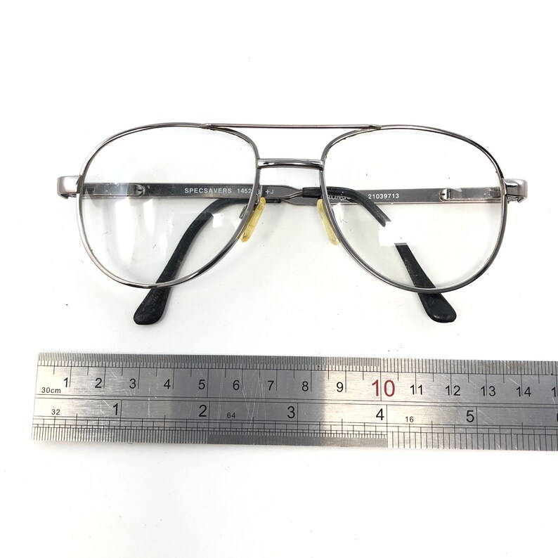 Peut inclure: Lunettes de vue de style aviateur avec une monture argent&eacute;e et des verres transparents. Les branches sont noires et il y a un double pont. Le texte "SPECSAVERS 1457" et "21039713" est visible. Une r&egrave;gle est en dessous.